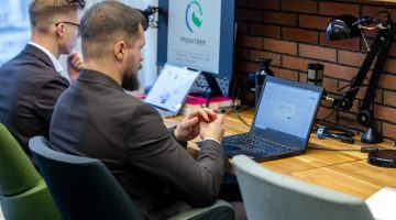 Dwóch mężczyzn w garniturach pracuje przy biurkach w biurze. Na stole leżą laptopy, jeden z nich otwarty z widocznym dokumentem. Na monitorze obok logo „Impactden”. Jednym z mężczyzn jest Karol Molendowski - autor tekstu na temat tego, Jak przygotować sprawozdanie finansowe organizacji pozarządowej.