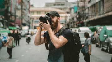 Na zdjęciu mężczyzna z profesjonalnym aparatem fotograficznym mie®zacy obiektywem w jakieś miejsce. W tle zgiłk miasta. Mężczyzna ma brodę, jest ubrany w t-shirt i czapkę z daszkiem.